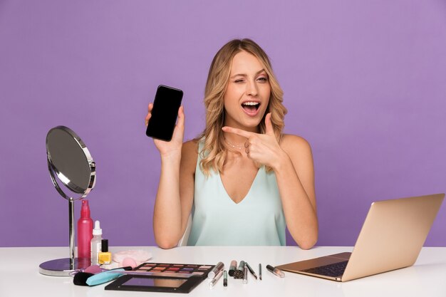 vrolijke positieve jonge vrouw met laptopcomputer en cosmetica met spiegel wijzend met weergave van mobiele telefoon.