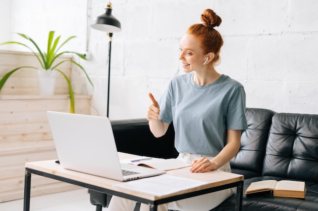 Vrolijke roodharige jonge vrouw praat over videogesprek en toont gebaar duim omhoog Succesvolle zakenvrouw freelancer heeft online briefing Concept van werken op afstand of onderwijs vanuit huis