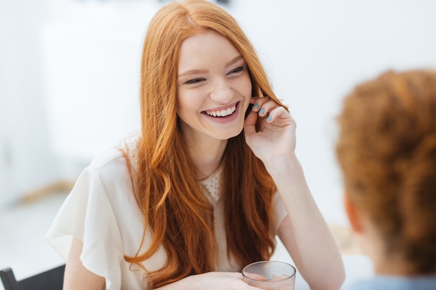 Vrolijke roodharige mooie jonge vrouw die zit en lacht met een vriend in café