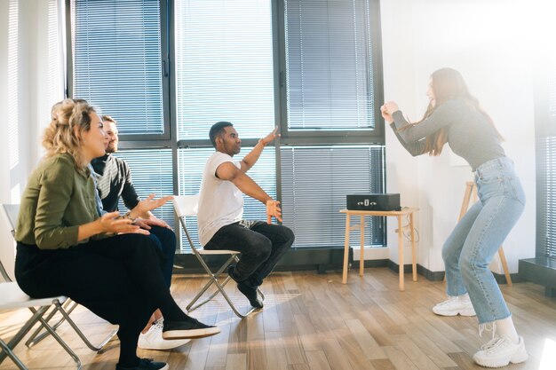 Vrolijke roodharige vrouw die charades speelt met vrienden die pantomime tonen bij het raam in het kantoor. groep activiteiten diverse multi-etnische collega's die in actieve spellen spelen tijdens teambuilding.