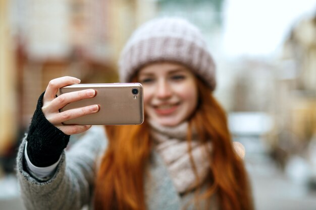 Vrolijke roodharige vrouw in winterkleding die selfie op haar mobiele telefoon neemt. Ruimte voor tekst