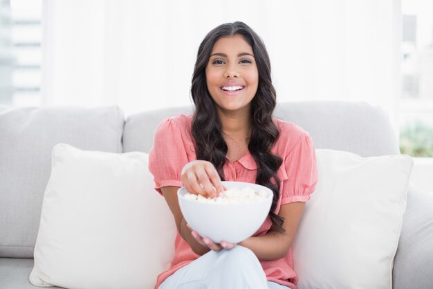 Vrolijke schattige brunette zittend op de bank holding popcorn kom