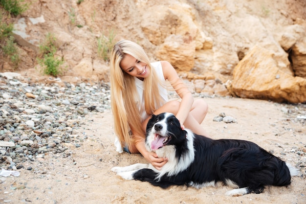 Vrolijke schattige jonge vrouw zittend met hond op het strand