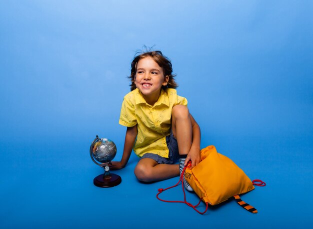 Vrolijke schooljongen zit op een blauwe achtergrond met een rugzak en een wereldbol. onderwijs concept