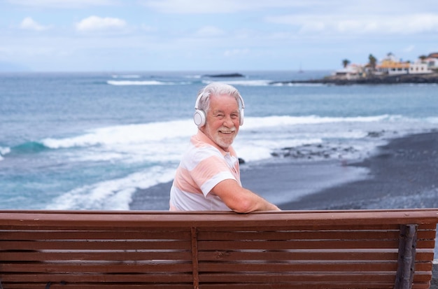 Vrolijke senior bebaarde man zittend op een bankje voor de zee ontspannen luisteren muziek door koptelefoon oudere man genieten van vrijheid en pensionering horizon over water