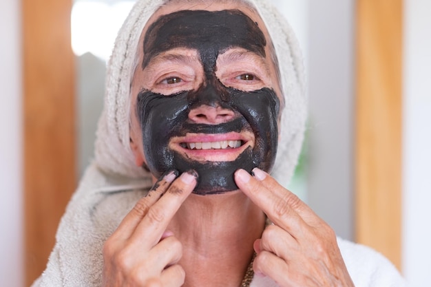 Vrolijke senior vrouw in badjas die een zelfgemaakt masker met detox-gezichtskool aanbrengt en voor de huid zorgt