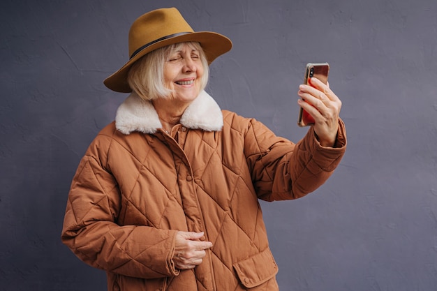 Vrolijke senior vrouw selfie te nemen op smartphone. Gelukkige oude dame in stijlvolle warme jas en muts