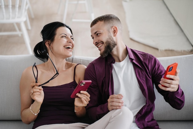 Vrolijke spaanse vrouw zit op de bank met echtgenoot genietend van vakanties met telefoon toothy glimlach Baard Blanke jonge man in violet shirt kijkt vriendin teder aan Vakantiepaar gezellig thuis