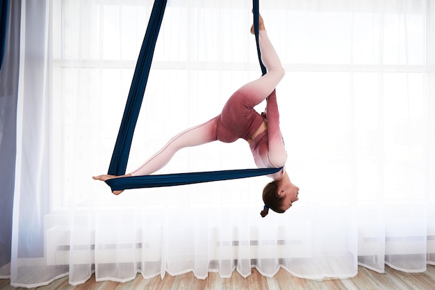 Vrolijke sportieve vrouw met top en legging die anti-zwaartekracht yoga-oefening uitvoert in een ruime gezondheidsclub met panoramische ramen.