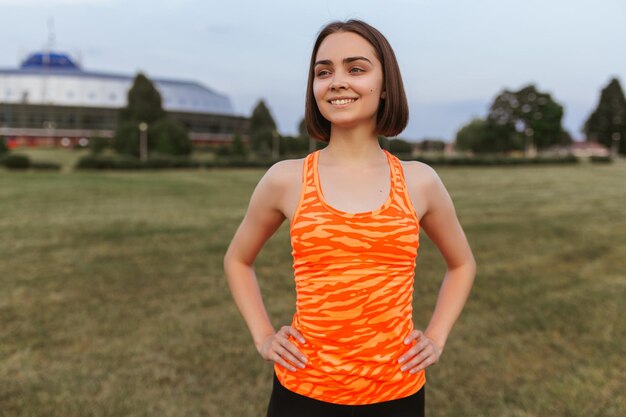 Vrolijke sportvrouw in oranje top met handen op taille in zomerpark tijdens training