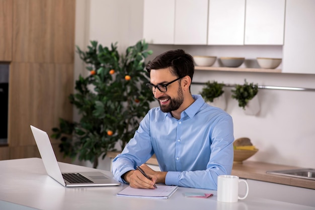 Vrolijke stemming Opgetogen lachende zakenman die aan tafel zit en aantekeningen maakt terwijl hij zijn laptop gebruikt