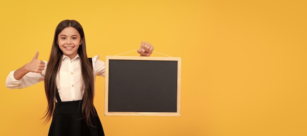 Vrolijke tiener meisje in uniform houdt schoolbord voor kopieerruimte met duim omhoog presenteren Banner van schoolmeisje student School kind leerling portret met kopie ruimte