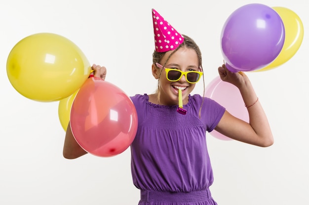 Vrolijke tienermeisje met ballonnen, in feestelijke hoed, blaast een pijp