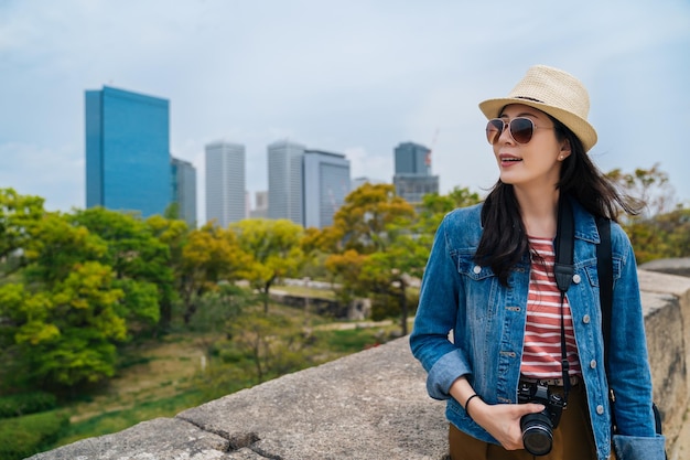 Vrolijke toerist met zonnebril met slr-camera die de stad in japan bezoekt. jonge aantrekkelijke reiziger geniet van de schoonheid van het groene park in de stad. dame die wacht om deel te nemen aan de kasteeltour van Osaka.