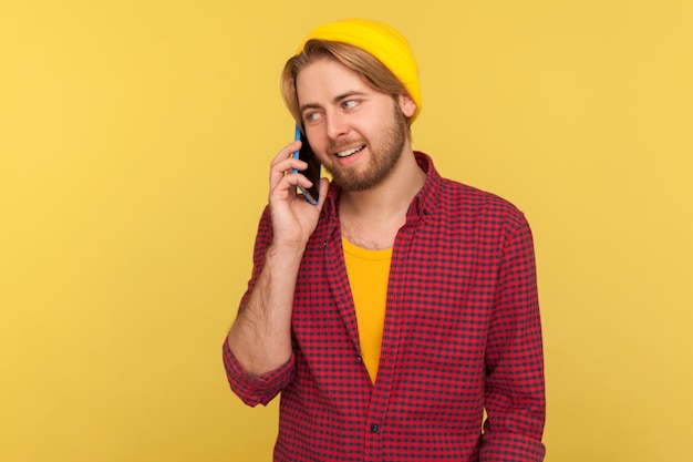 Vrolijke trendy man in hipster beanie muts en geruit hemd bellen, glimlachen tijdens het praten over de mobiele telefoon, een positief gesprek voeren over de telefoon. indoor studio-opname geïsoleerd op gele achtergrond