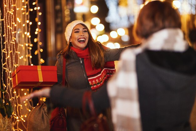 Vrolijke twee jonge vrouwen hebben plezier in de stadsstraat tijdens de kerstnacht. Ze lachen en geven cadeautjes aan elkaar.
