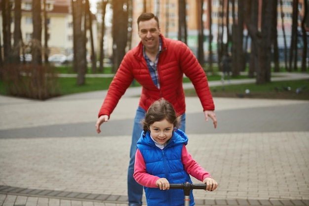 Vrolijke vader geniet van zijn vrije dag brengt tijd door met zijn kind, schattig klein meisje dat op een duwscooter rijdt in een stadspark Gelukkig familierelatiesconcept