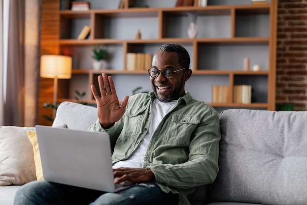 Vrolijke volwassen afro-amerikaanse man in glazen en casual zwaaiende hand en kijken naar computer