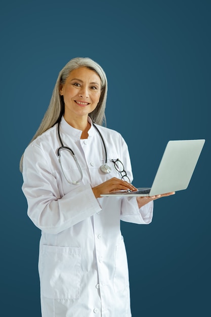 Vrolijke volwassen dame arts in wit gewaad maakt gebruik van moderne laptop staande op blauwe achtergrond