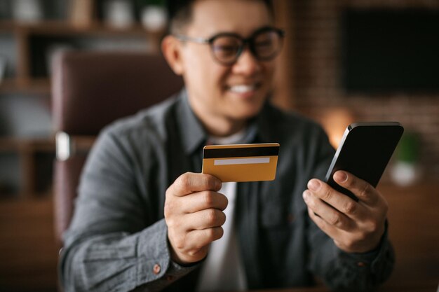 Vrolijke volwassen japanse man met een bril die op de telefoon typt, laat zien dat creditcard online winkelt, geïnvesteerd geld om op te starten