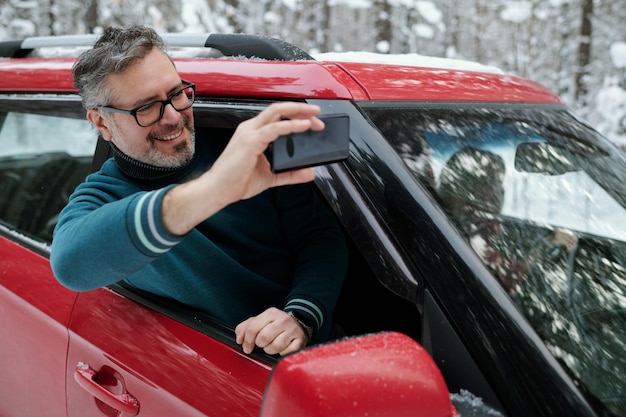 Vrolijke volwassen man die selfie neemt terwijl hij uit het autoraam stapt