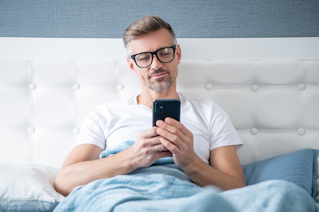 Vrolijke volwassen man in bed chatten op de telefoon met een bril op