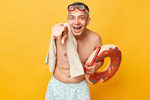 Vrolijke volwassen man met korte zwembroek zwembroek snorkelbril met donut rubberen ring en tas geïsoleerd op gele achtergrond lachend wijzend naar camera die jou de schuld geeft