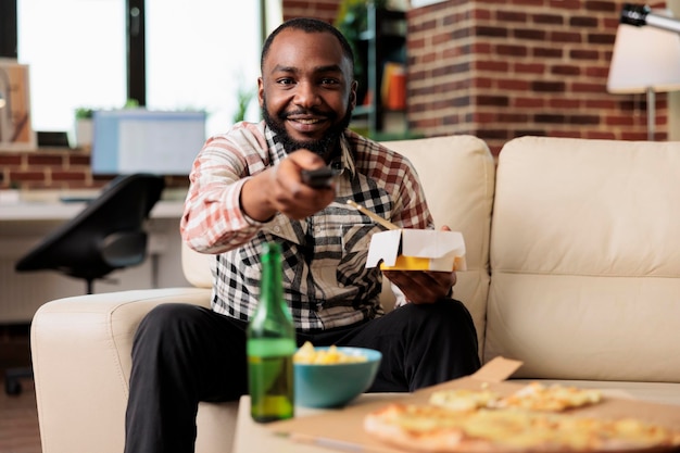 Vrolijke volwassen schakelende kanalen met tv-afstandsbediening en noedels eten van afhaalmaaltijden op de bank. Plezier hebben met film of film, eetstokjes gebruiken om afhaalmaaltijden te eten.