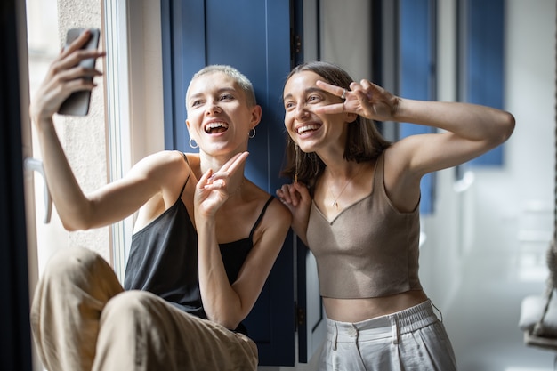 Vrolijke vriendinnen of lesbisch koppel hebben plezier, maken selfie-foto's of praten online via de mobiele telefoon die binnenshuis bij het raam zit