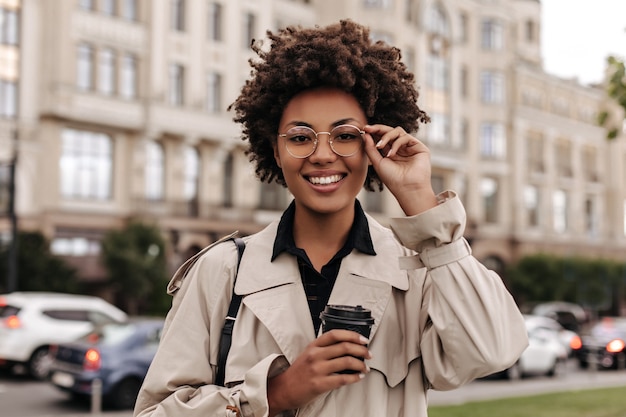 Vrolijke, vrolijke brunette krullende vrouw in beige trenchcoat zet een ronde bril op en houdt een koffiekopje buiten