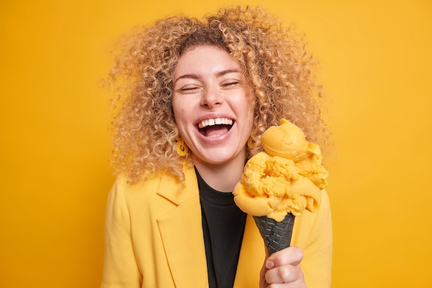 Vrolijke vrolijke jonge vrouw heeft krullend borstelig haar lacht breeduit als ze iets grappigs hoort trakteert zichzelf met lekker koud ijs, elegant gekleed poseert tegen de gele muur.