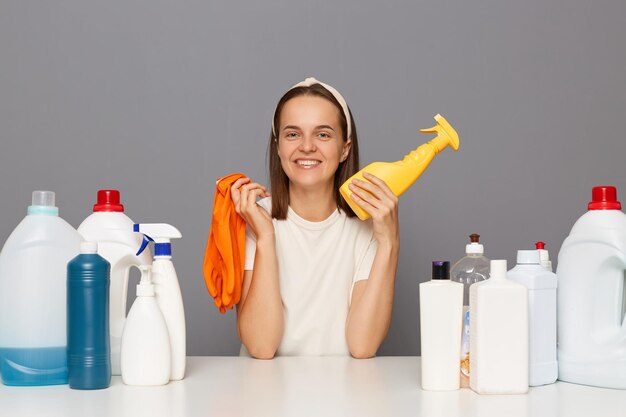 Vrolijke vrolijke vrouw die zich voordeed op de werkplek met schoonmaakmiddelen geïsoleerd over grijze achtergrond met rubberen handschoenen kijkend naar de camera met een blij gezicht