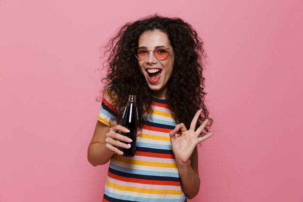 vrolijke vrouw 20s met krullend haar frisdrank drinken uit glazen fles geïsoleerd op roze