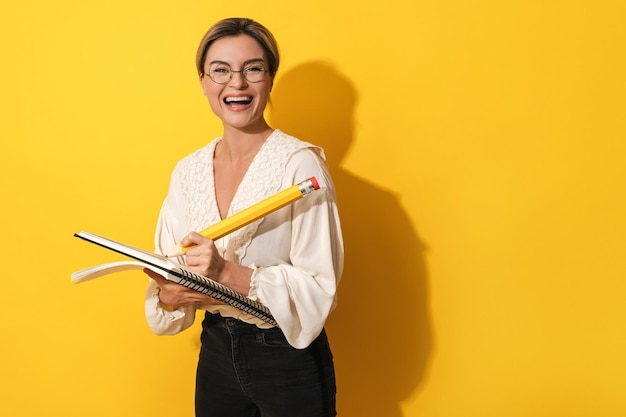 Vrolijke vrouw die een bril draagt met een groot potlood en een notitieboekje op een gele achtergrond