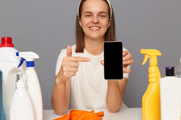 Vrolijke vrouw die een wit t-shirt draagt en omringd wordt door het schoonmaken van chemische benodigdheden wijzend op een smartphone met een leeg display-advertentiegebied dat geïsoleerd over een grijze achtergrond zit