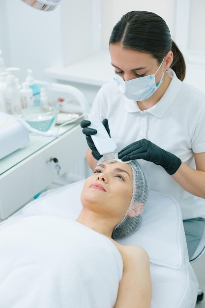 Vrolijke vrouw die lacht terwijl een geconcentreerde schoonheidsspecialist een ultrasone reinigingsprocedure op haar voorhoofd uitvoert