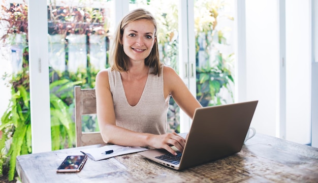 Vrolijke vrouw die met laptop aan het keukenbureau werkt en naar de camera lacht tegen het raam van de villa