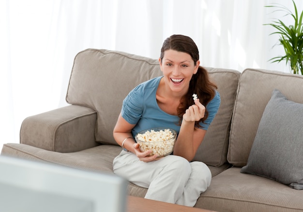 Vrolijke vrouw die popgraan eet terwijl het letten van een op film op televisie