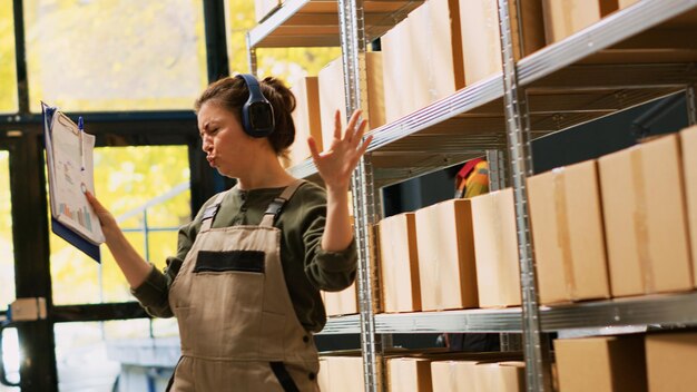 Vrolijke vrouw die ronddanst in het depot, luistert naar muziek en controleert de logistieke voorraad vóór distributie. Vrouwelijke manager die plezier heeft met dansbewegingen en merchandisepakketten beoordeelt. Uit de hand geschoten.