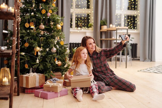 Vrolijke vrouw en schattig klein meisje in pyjama selfie maken door versierde kerstboom in het midden van de huiskamer