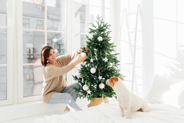 Vrolijke vrouw in comfortabele kleding siert kerstboom in moderne ruime slaapkamer kijkt met een glimlach naar rashond zittend op bed groot raam achter Nieuwjaar tijd vakantie voorbereiding