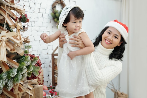 Vrolijke vrouw in kerstmuts die dochtertje optilt wanneer ze poseert naast de versierde kerstboom bij ...