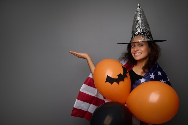 Vrolijke vrouw in tovenaarshoed, gewikkeld in Amerikaanse vlag, houdt oranje luchtballen vast, glimlacht brede glimlach kijkend naar camera, tilt haar handpalm op, houdt denkbeeldige kopieerruimte vast voor advertentie op grijze achtergrond