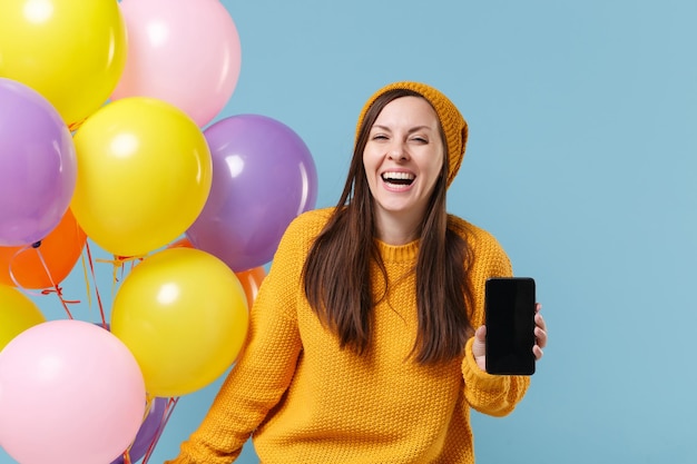 Vrolijke vrouw in trui hoed poseren geïsoleerd op blauwe achtergrond. verjaardag vakantie partij mensen emotie concept. bespotten kopie ruimte. vieren houd luchtballonnen mobiele telefoon met leeg leeg scherm.