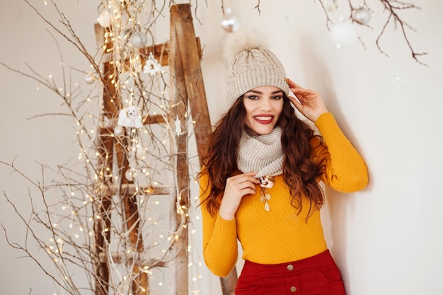 Vrolijke vrouw in warme muts en sjaal, gele trui en rode rok poseert naast kerstversiering.