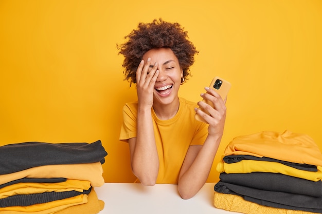 Vrolijke vrouw met Afro-haar maakt gezicht palm glimlacht breed houdt mobiele telefoon voelt zich blij gekleed in casual t-shirt omringd door twee stapels gevouwen kleding geïsoleerd over geel