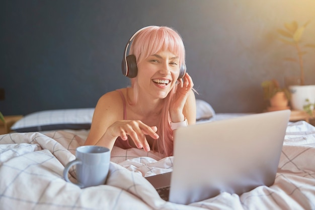 Vrolijke vrouw met koptelefoon luistert naar muziek op laptop op bed