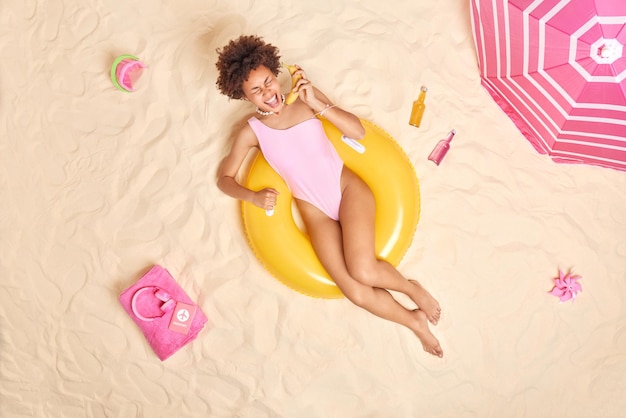 Vrolijke vrouw met krullend haar gekleed in zwembroek houdt banaan in de buurt van oor doet alsof ze een telefoongesprek heeft ligt in de zon op het strand poses op opgeblazen zwemmen roept luid geniet van zomervakantie