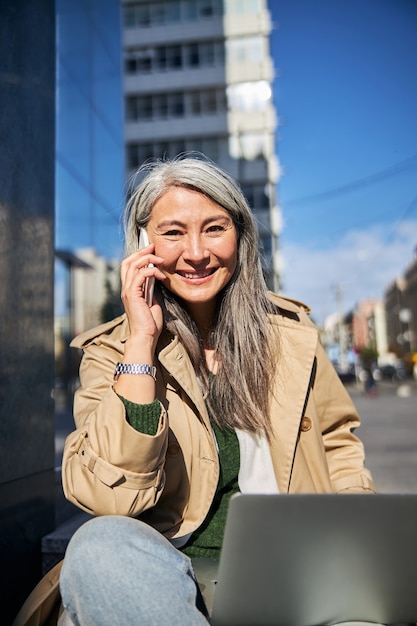 Vrolijke vrouw met laptop praten op mobiele telefoon buitenshuis