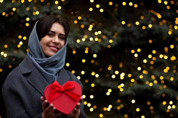 Vrolijke vrouw met rode geschenkdoos in de vorm van een hart op de achtergrond van wazig bokeh lichten. Lege ruimte